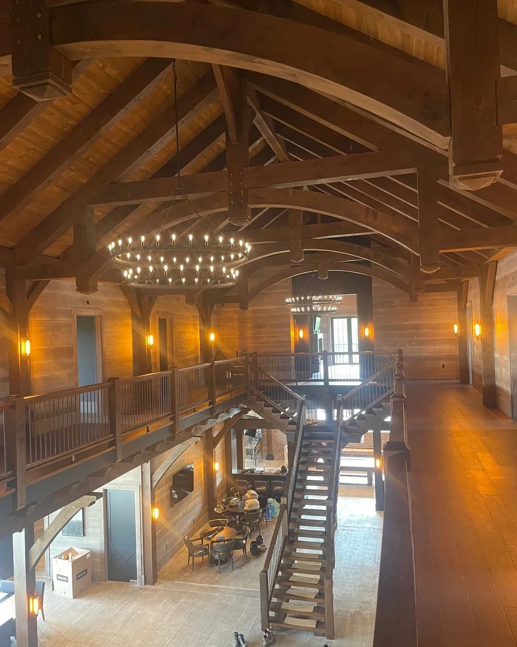 A timber frame great room with an exposed truss roof and a large circular chandelier over a staircase.