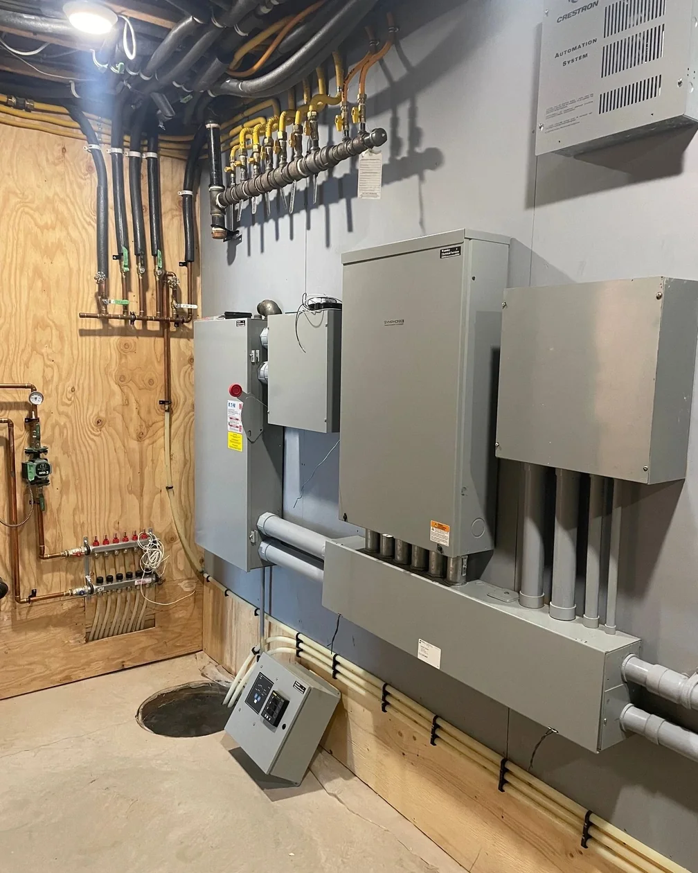A mechanical room wall with a row of grey electrical panels, conduit runs and a manifold.