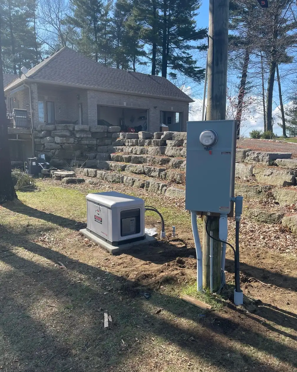 A standby generator on a concrete pad beside a rural house, with a meter and transfer switch mounted on a pole.