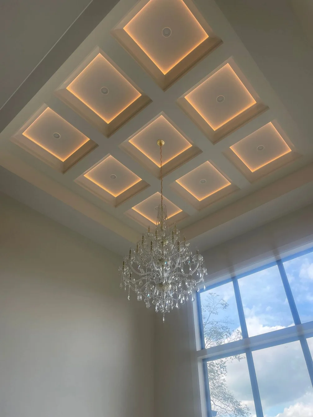 A coffered ceiling with each recessed panel edge lit, and a crystal chandelier alongside a tall window.