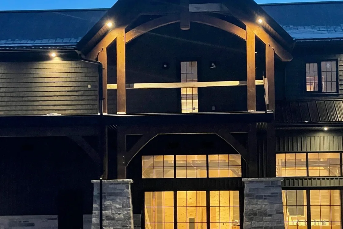 A large timber frame house at dusk with warm light coming through tall windows and under the eaves.