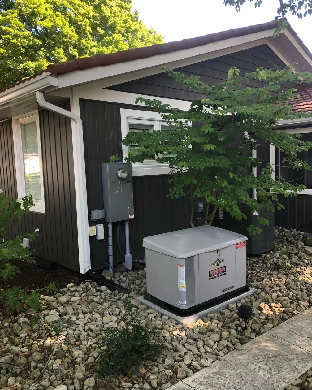 A standby generator on a gravel bed against the cedar clad wall of a house, with the meter and disconnect above it.