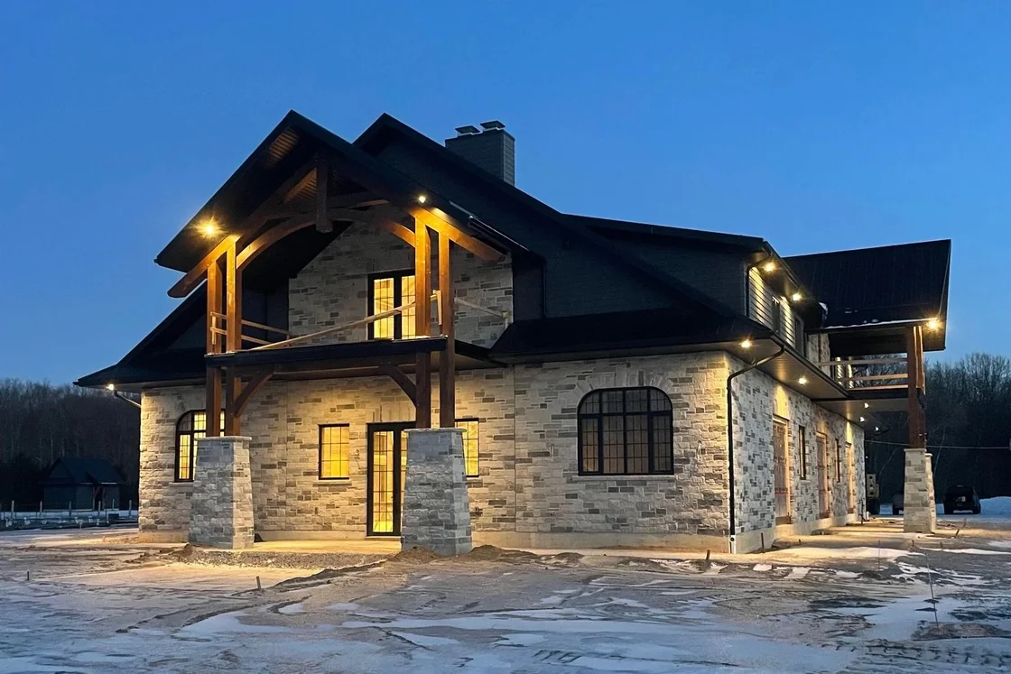 A stone and timber house at blue hour with warm light glowing from the windows and under the eaves, snow on the ground.