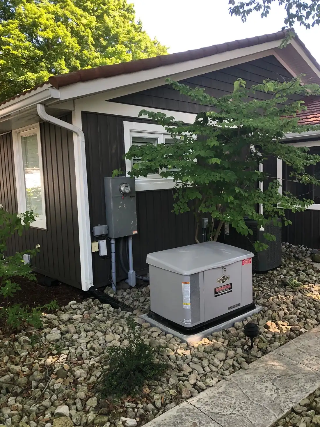 A standby generator on a gravel bed against the cedar clad wall of a house, with the meter and disconnect above it.