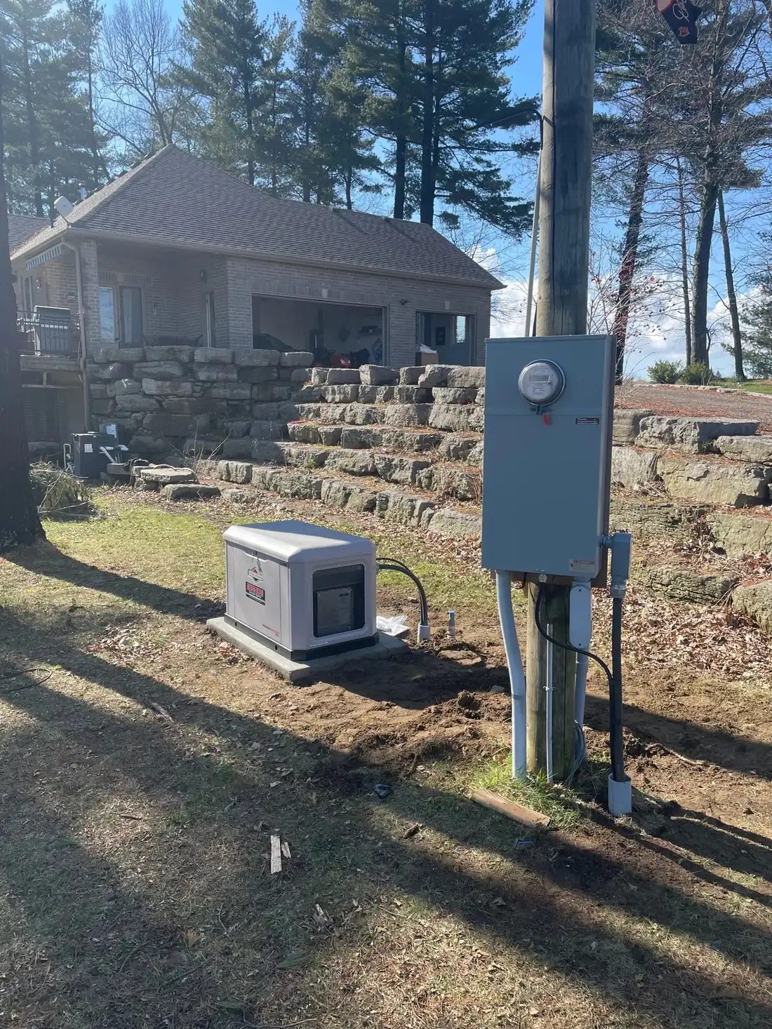 A standby generator beside a rural house with a meter and transfer switch on a pole.