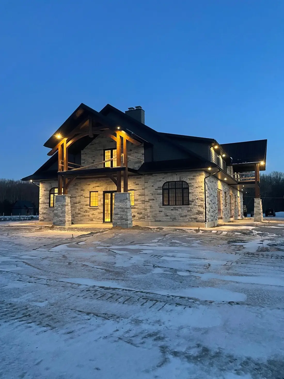 A stone and timber house at blue hour with warm light glowing from the windows and under the eaves, snow on the ground.